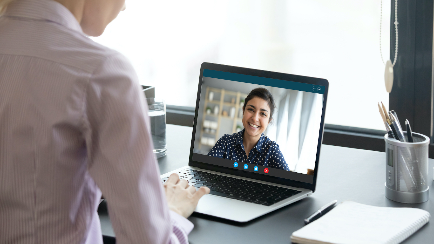 Indian girl communicate with friend on-line by video call, pc screen view over female shoulder. Mental health expert online therapy, colleagues work on common project use videoconferencing app concept