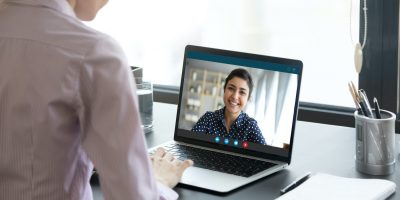 Indian girl communicate with friend on-line by video call, pc screen view over female shoulder. Mental health expert online therapy, colleagues work on common project use videoconferencing app concept